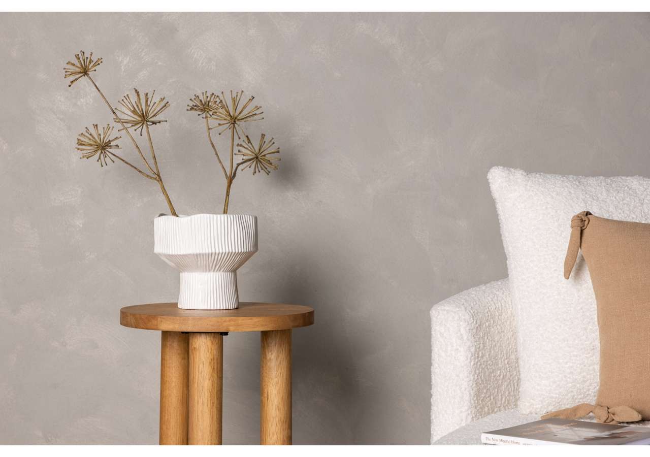 Ljus inredningsdetalj med vit blomvas och sidenväxter på träbord, modern sängstopp med beige kudde.