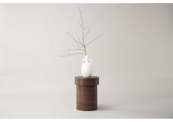 Enkel vit vas på trästub med gren mot ljus bakgrund.
