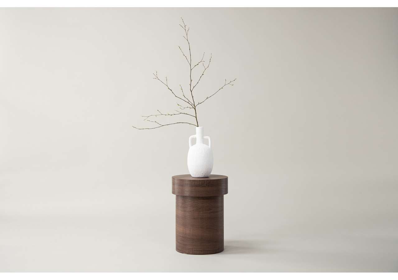 Enkel vit vas på trästub med gren mot ljus bakgrund.