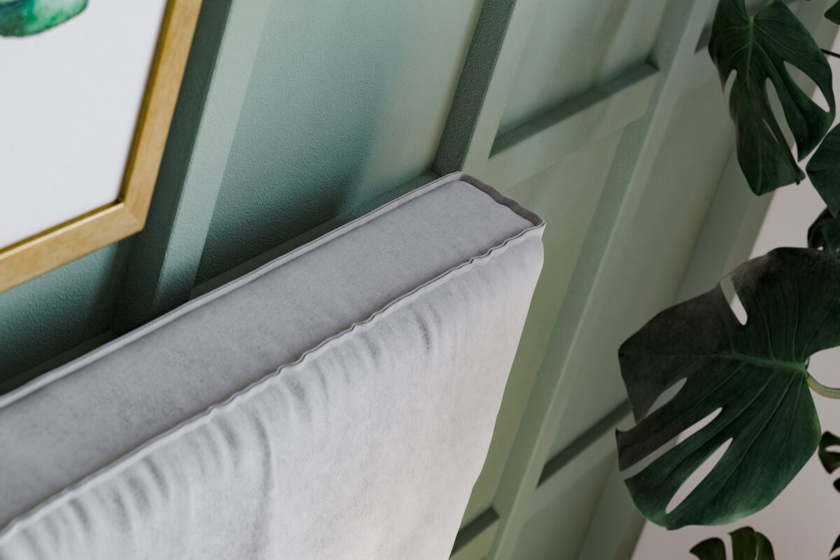 Sängram i ljusgrå sammet med grön vägg och växt i bakgrunden.