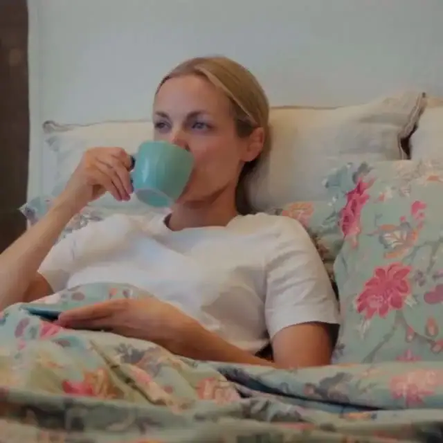 Avslappnad kvinna i säng med vackra sängtäcken, njuter av morgonkaffe och vila.