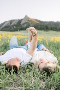 Couple laying in field of wildflowers with arms up looking at her engagement ring