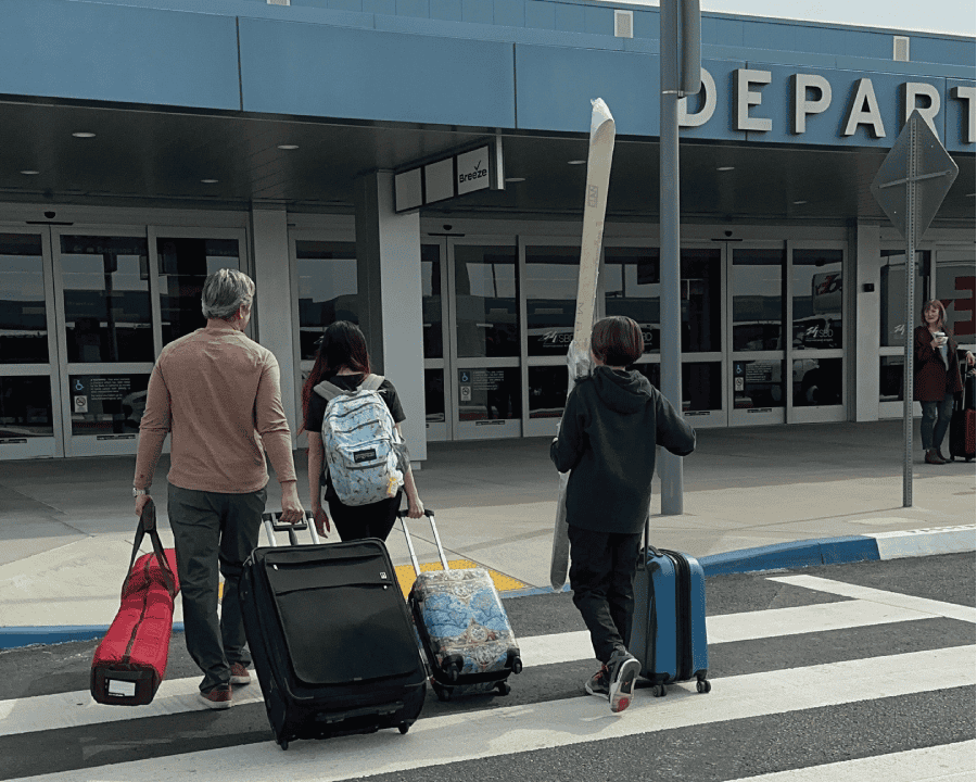 A father, son and daughter walking to the San Bernardino International Airport terminal with luggage and snow skis. A woman in standing at the curb with luggage and a cup of coffee.