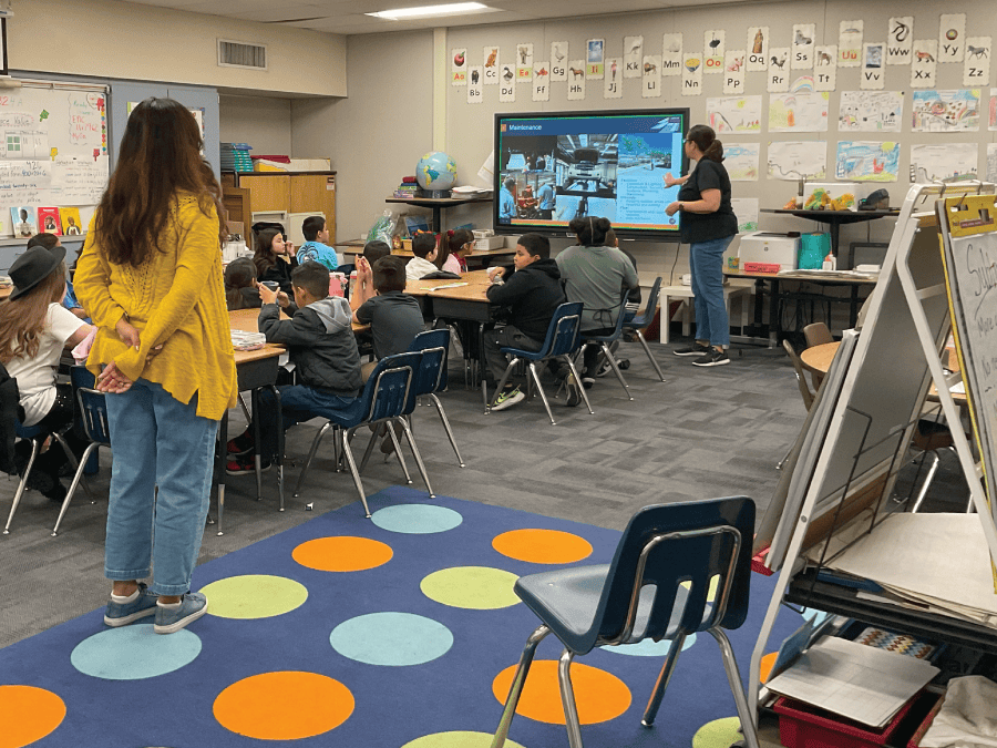 A woman standing at the front of a classroom full of children color pointing to the TV with an airport presentation showing with the teacher standing in the background