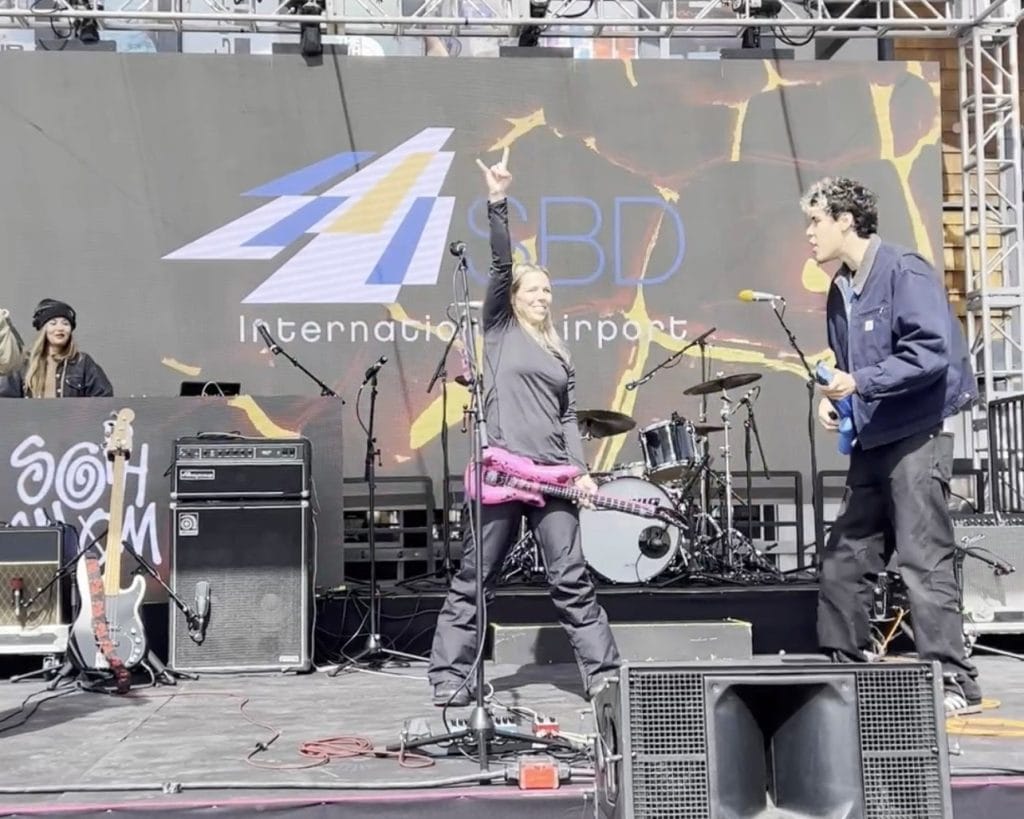 Woman standing on a concert stage with arm raised in the air with excitement, holding an inflatable guitar while participating in an air guitar contest. SBD logo in background.
