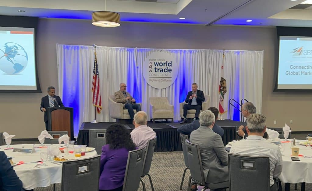 Two men seating on a stage with a moderator at a podium presenting to an audience