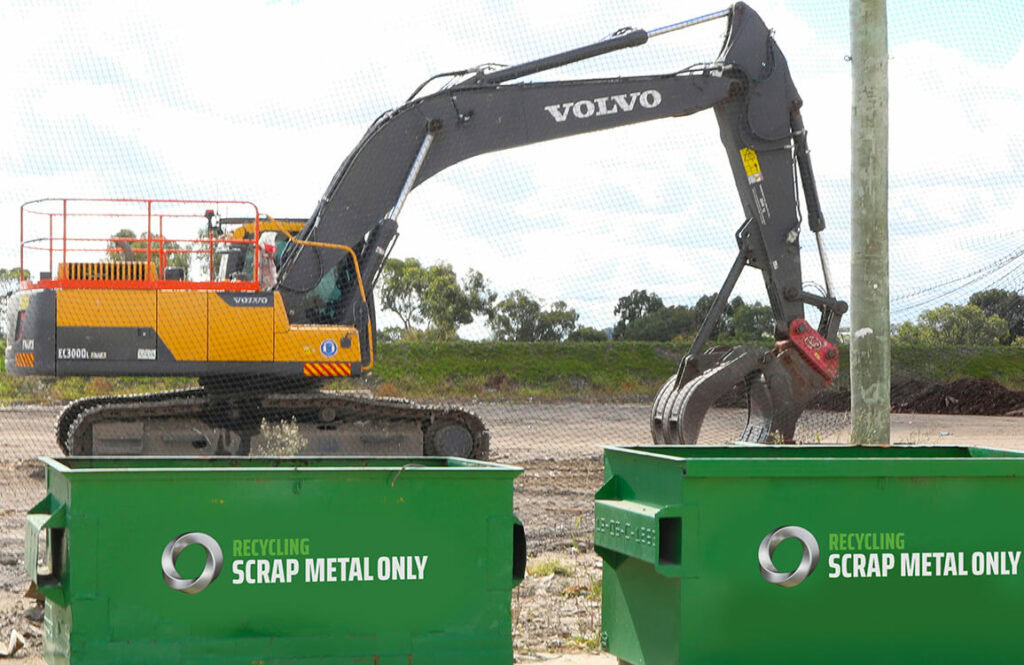 Heavy-duty excavator at scrap metal recycling site with green bins.