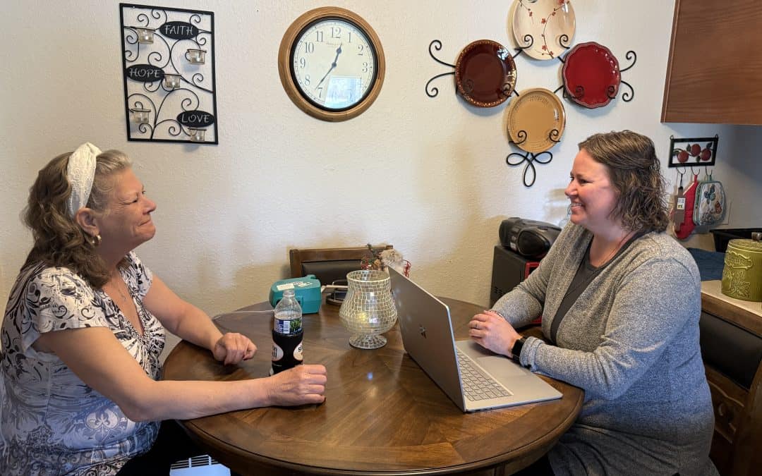 Elderly woman and caregiver talking at a kitchen table with laptop and water bottle.
