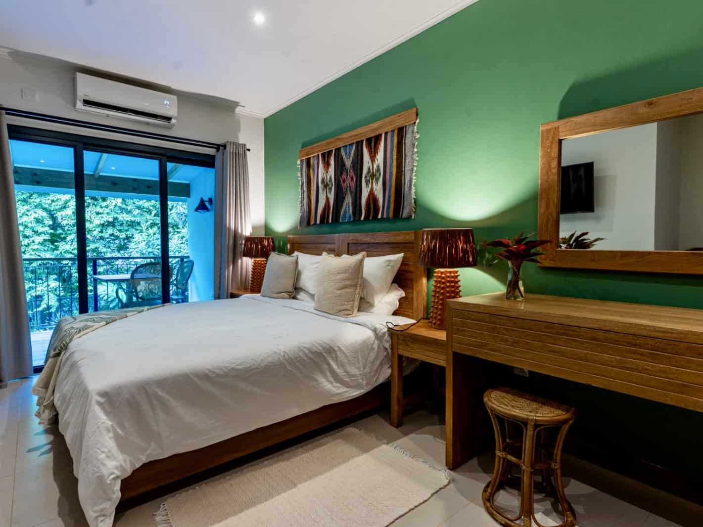 A comfortable hotel room with a large bed, green accent wall, wooden furniture, and a balcony overlooking lush Seychelles foliage.