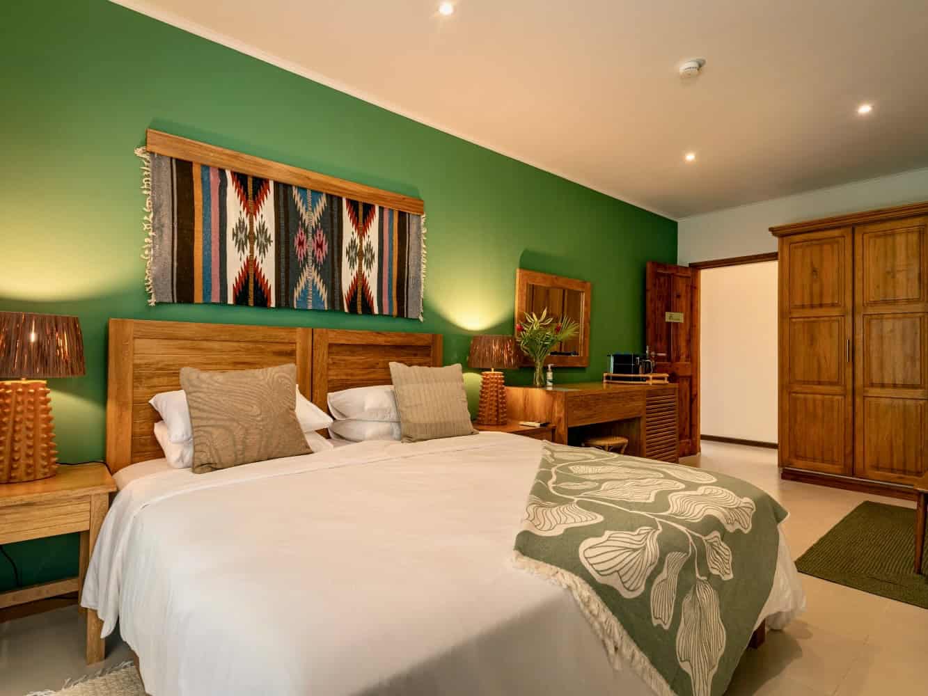 Lit dans une chambre avec mur vert, décoration en bois naturel, linge blanc, coussins beige, lampe en bois, et ambiance chaleureuse.