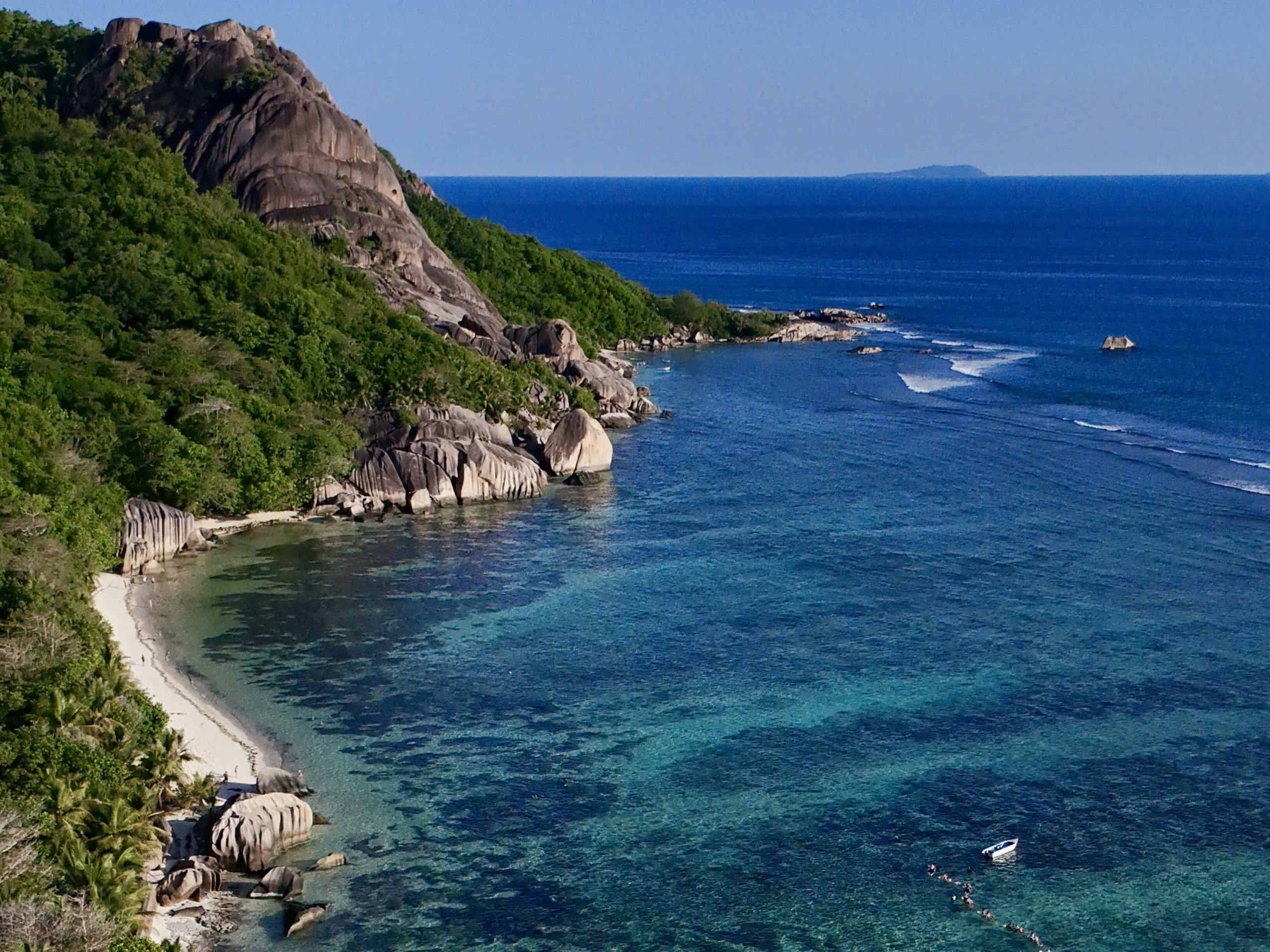 Belles falaises rocheuses et eaux turquoise de la plage de Mahé, Seychelles, avec des kayaks en mer et une végétation luxuriante en arrière-plan.
