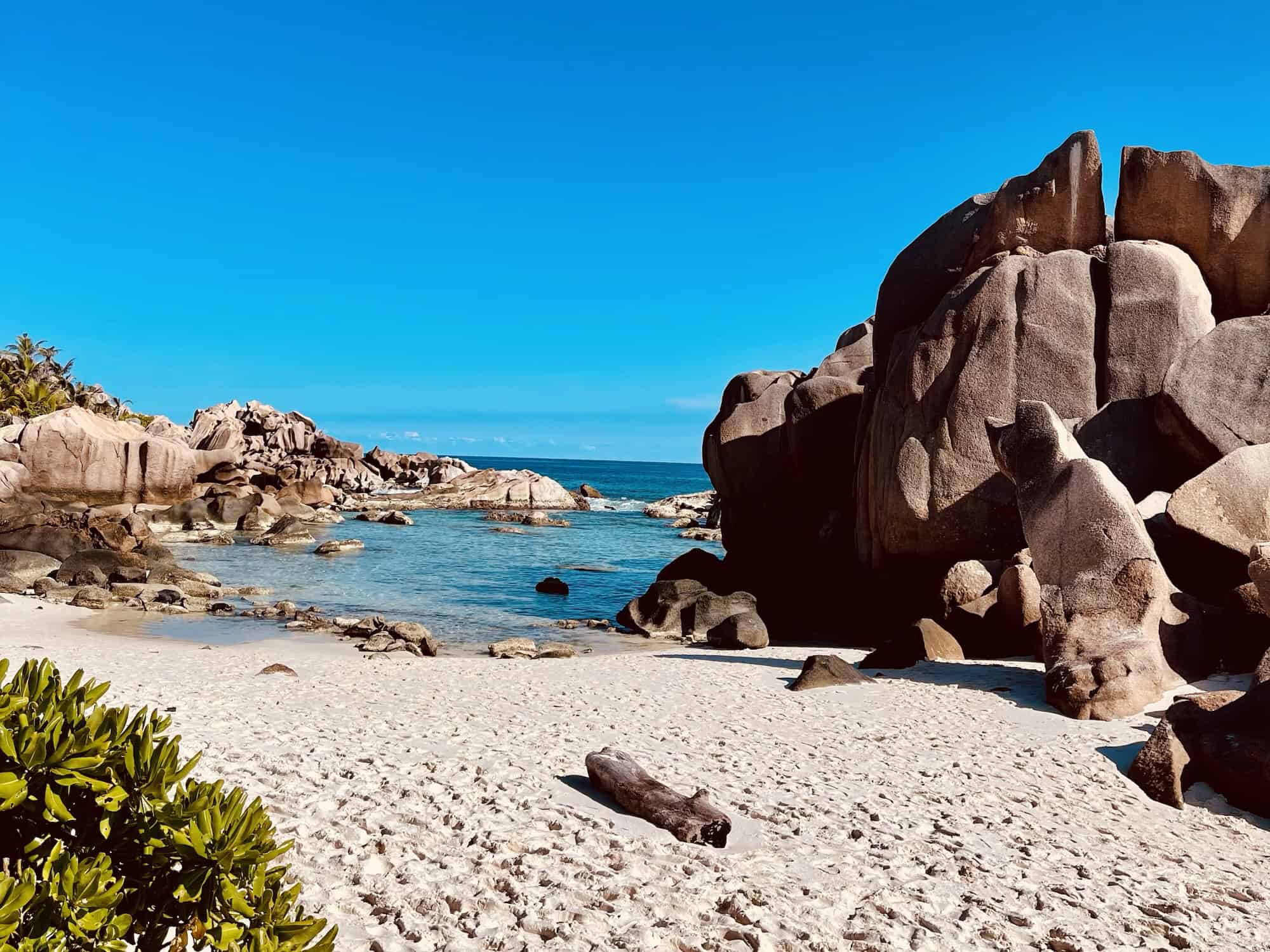 Hauteurs de rochers granitiques sur une plage de sable blanc à Mahé, Seychelles, Mer turquoise, ciel bleu, végétation tropicale dense en arrière-plan.