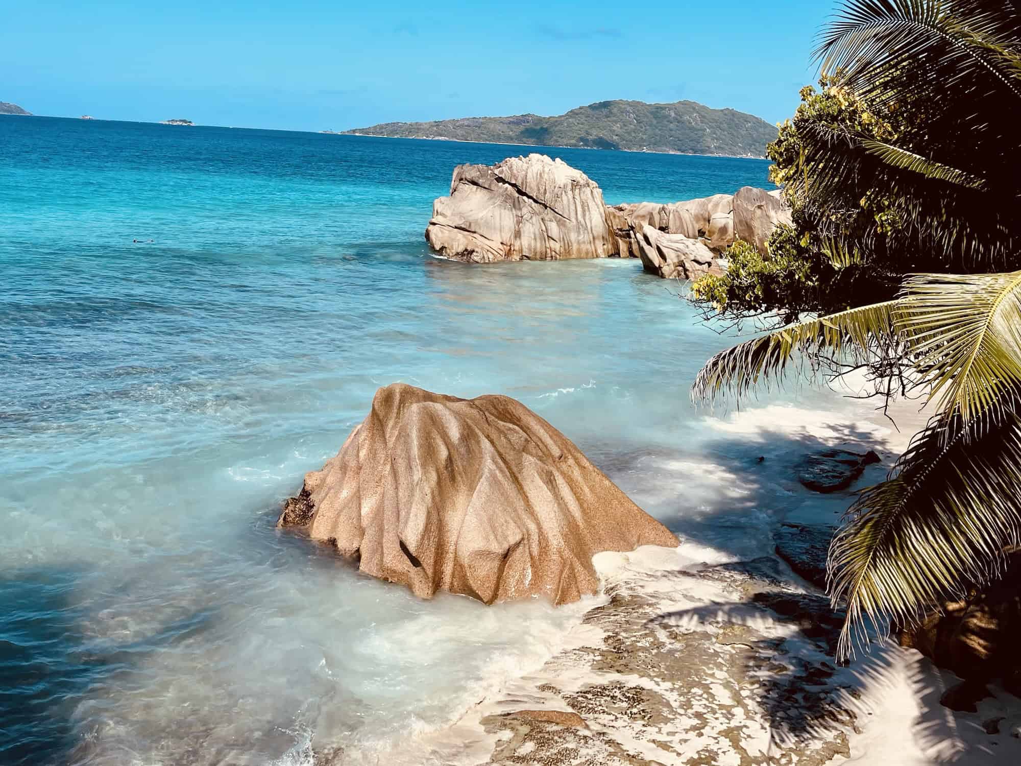 Rocks près d'une plage de Seychelles avec palmiers, eau turquoise et ciel bleu.