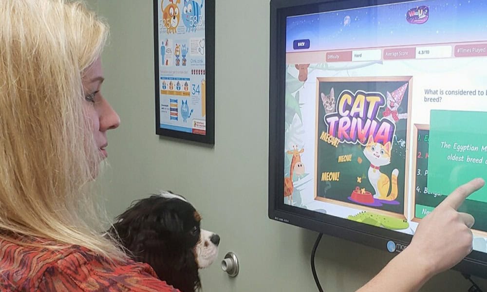 A veterinarian pointing at a computer screen displaying "cat trivia" to a small dog in her lap, in an office setting.