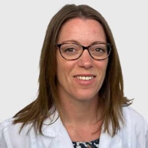 Professional headshot of a smiling veterinarian with long brown hair wearing glasses and a white lab coat over a patterned blouse, against a plain, light background.