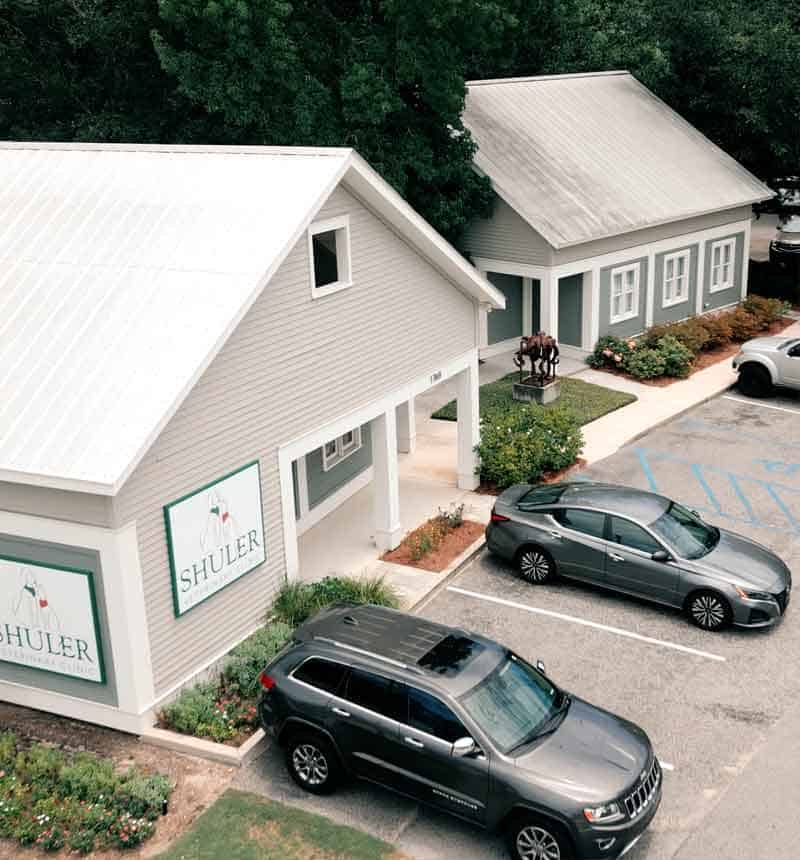 Aerial view of two gray buildings with white trim, featuring signs that say "Shuler." Two parked cars are visible in front, alongside a small garden area, under overcast skies.