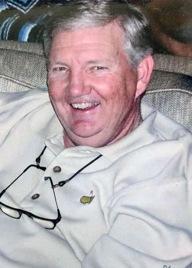 Smiling middle-aged veterinarian sitting comfortably on a couch, wearing a white shirt with a small colorful logo on the pocket, and posing for the camera.