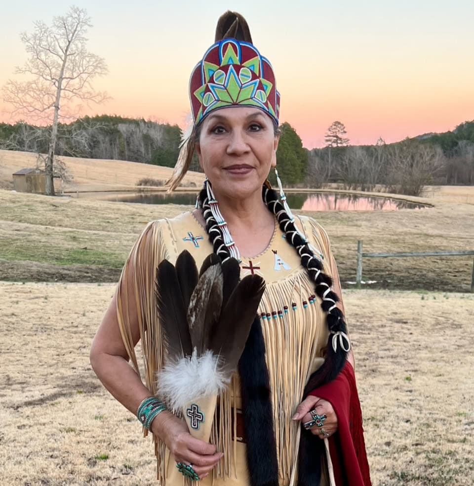 Protective Native American woman in ceremony attire, holding feathers outdoors at sunset.