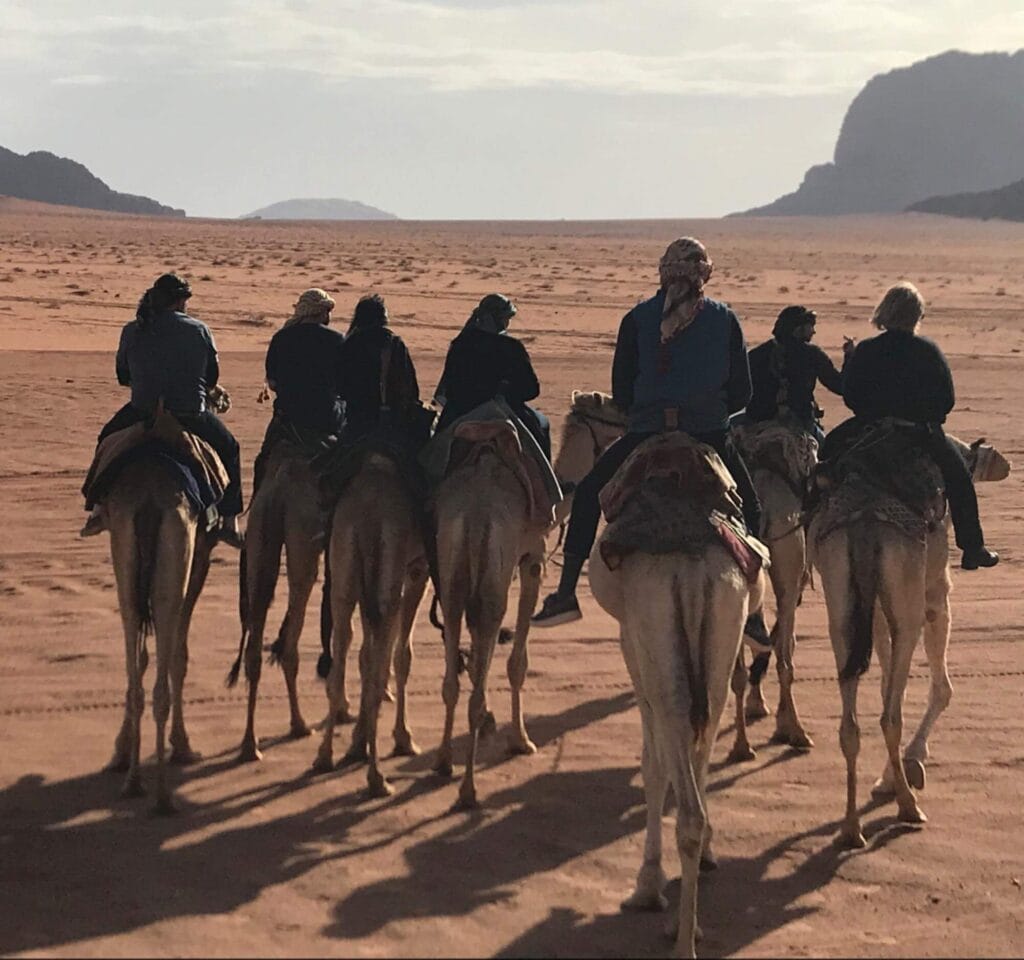 People riding camels across a vast desert landscape during sunset.