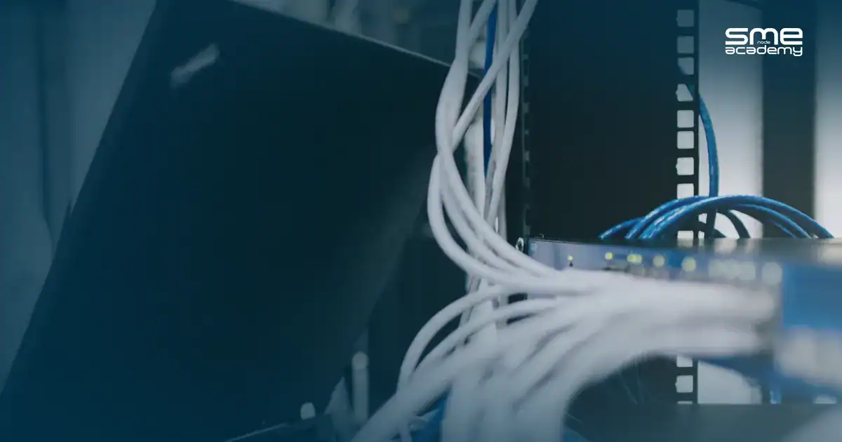 A network engineer's open laptop beside a patch panel of white ethernet cabling, graded with the SMEnode Academy navy brand tint.