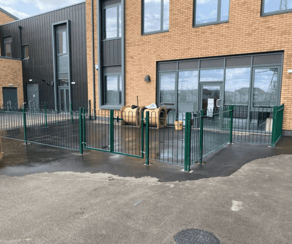 Green metal fence enclosing small outdoor area with yellow play equipment in front of brick building