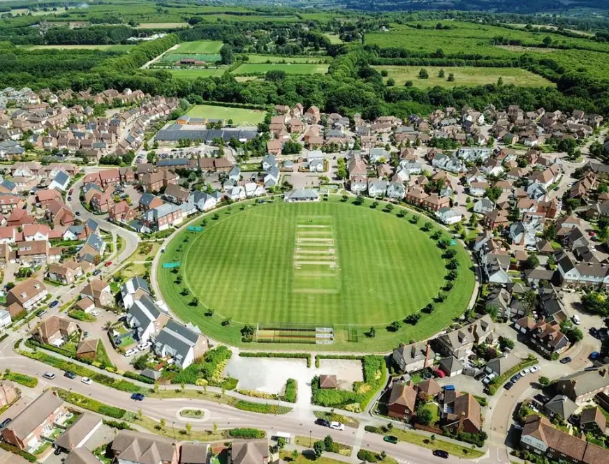 Aerial view of a lush green sports field surrounded by a suburban neighborhood with houses. Ideal for cricket, football, or community events, showcasing eco-friendly urban recreation spaces.