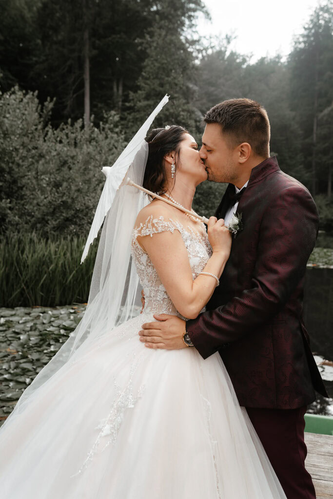 Romantisches Hochzeitsfoto eines Paares in Frankfurt, das sich liebevoll küsst, mit Regenschirm und Natur im Hintergrund, aufgenommen von einem professionellen Hochzeitsfotografen.