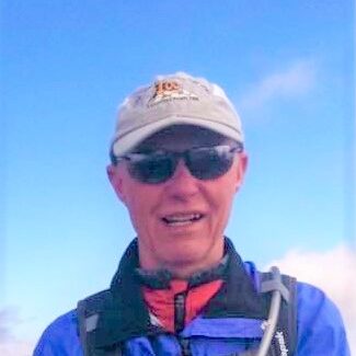 A male trail runner wearing sunglasses and a cap smiling outdoors against a blue sky during a running event.