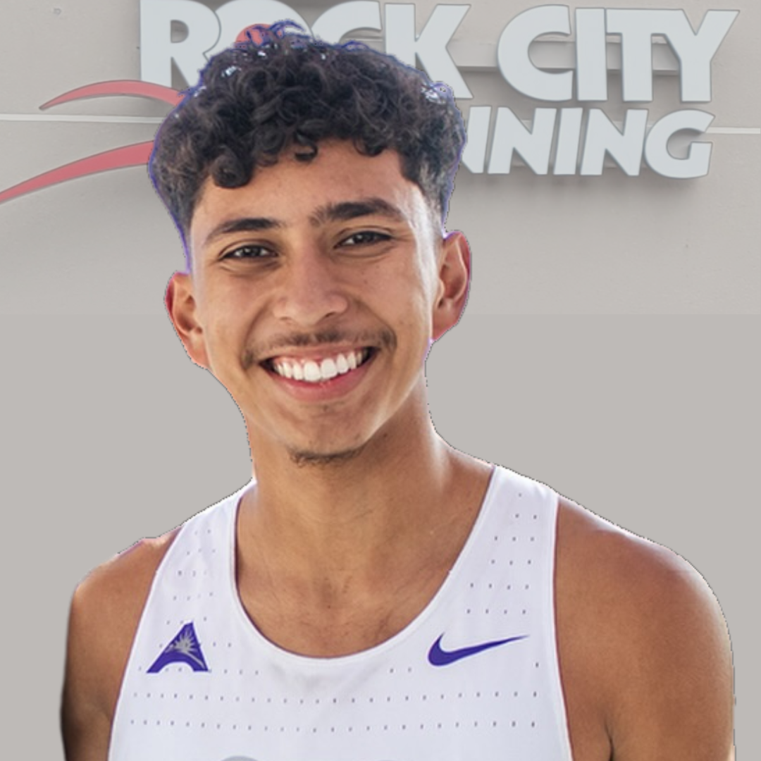 Young male runner smiling, wearing sports gear in front of Rock City Running store in Little Rock, Arkansas, showcasing athletic apparel and running shoes.