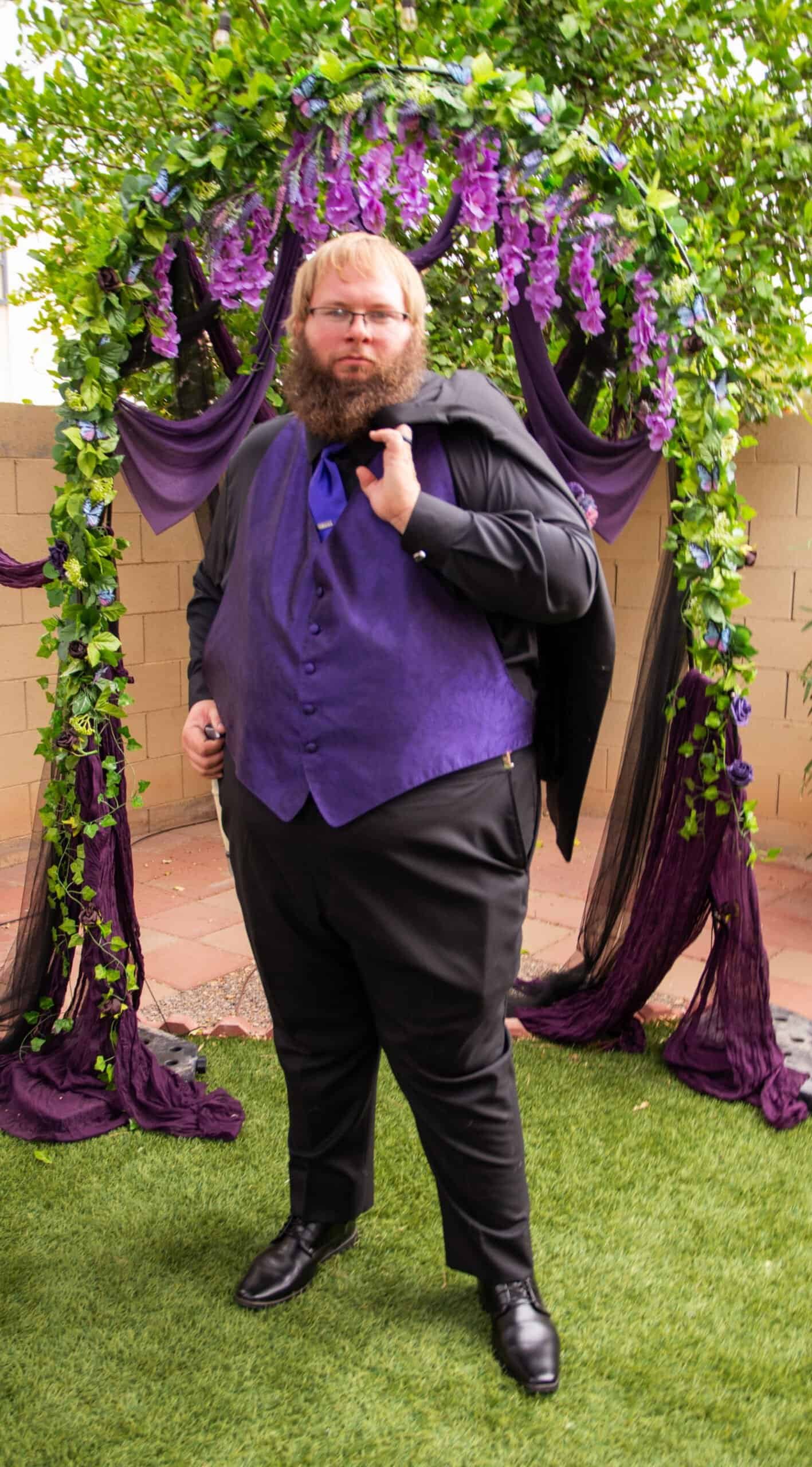 A man dressed in formal black suit and purple vest posing at a wedding ceremony with floral decorations.