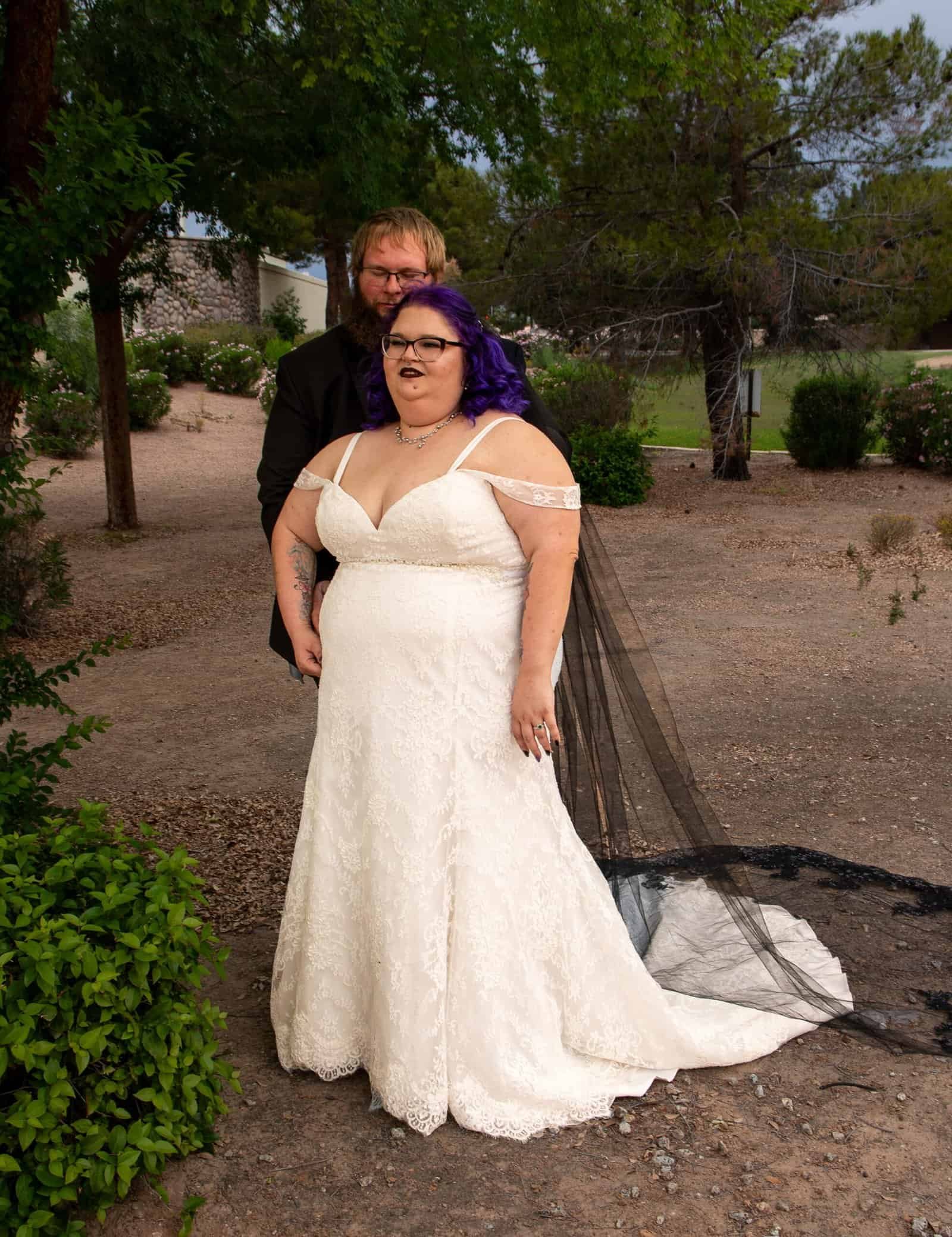A bride and groom pose together outdoors during their wedding, surrounded by trees and natural scenery.