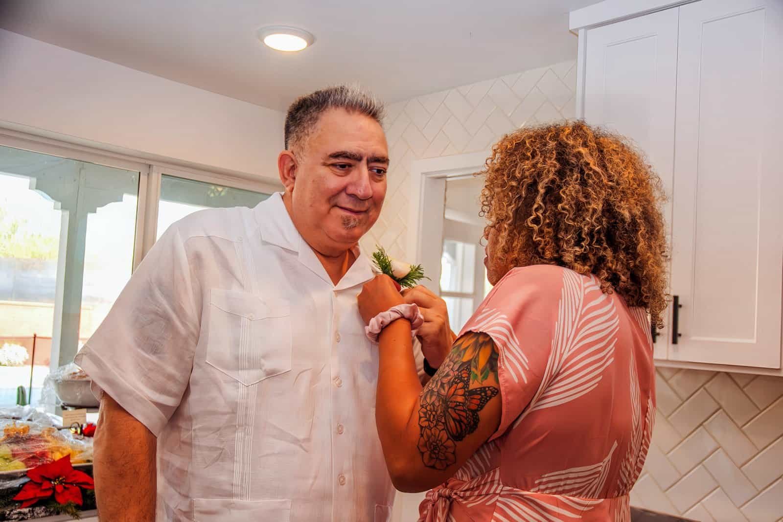 A woman with curly hair and tattoos pins a boutonniere on a smiling man in a bright kitchen setting, capturing a special event.