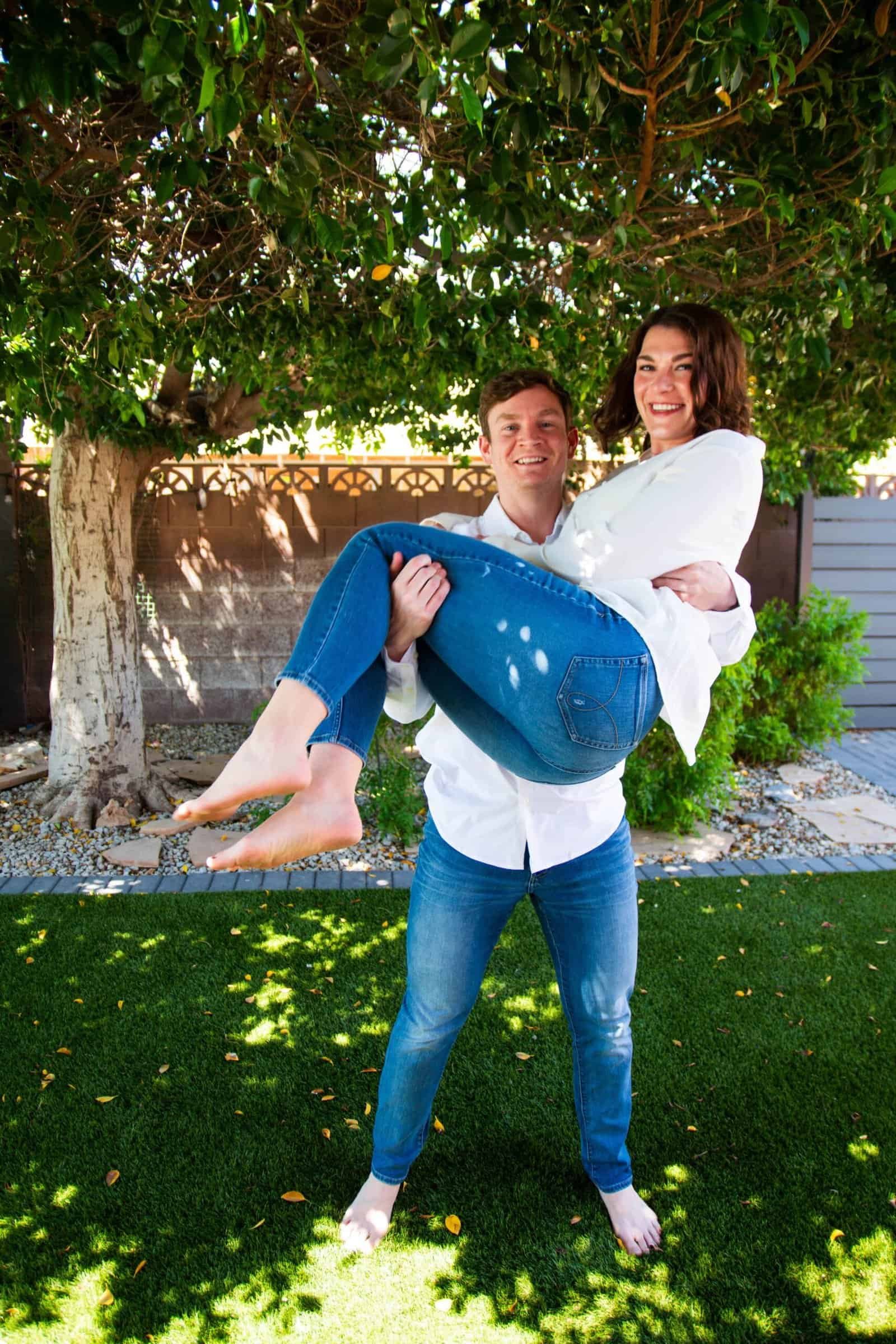 A man lifts a woman in his arms under a large tree, smiling and having fun in a sunny backyard setting.