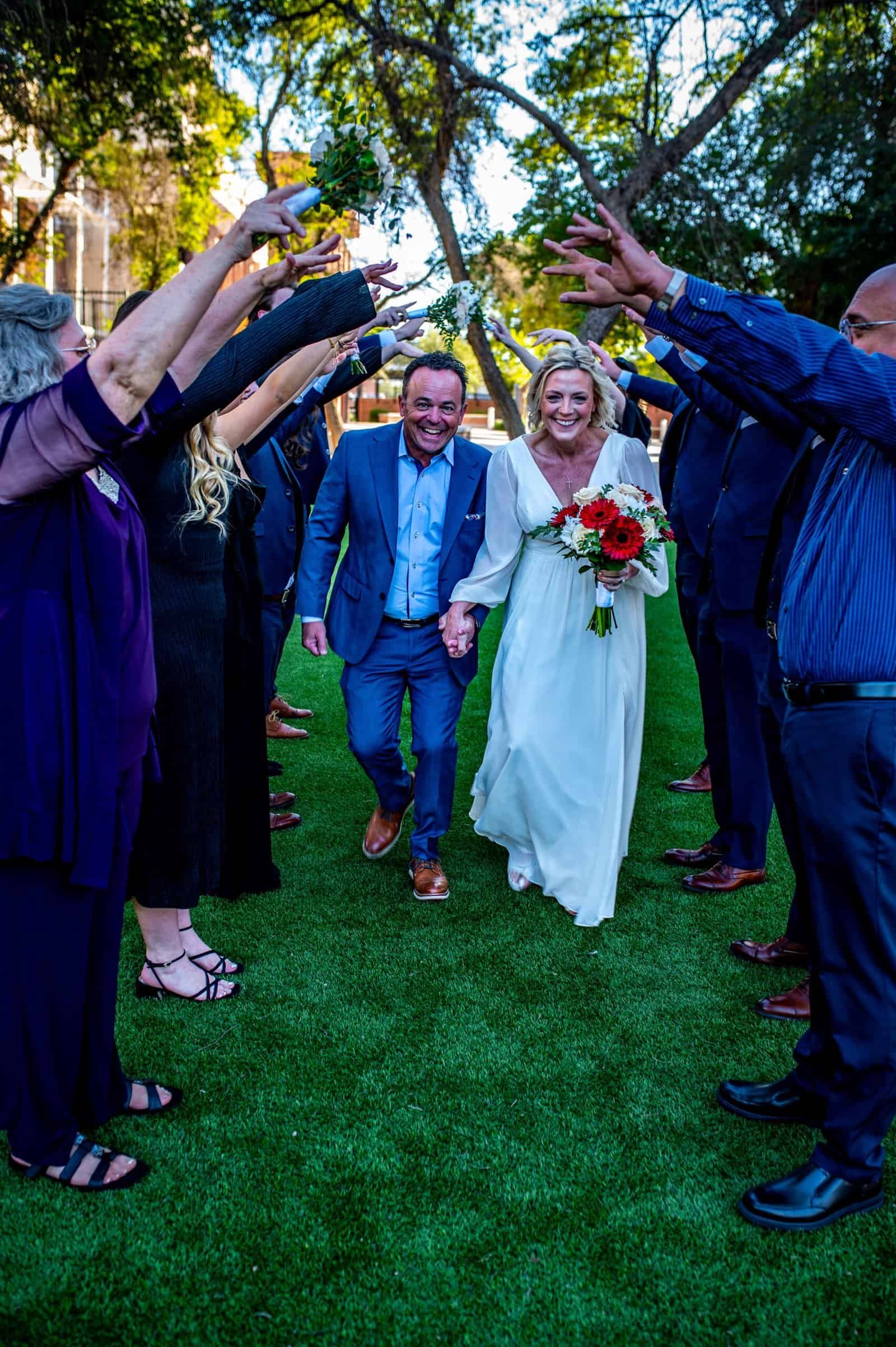 A joyful wedding moment where the bride and groom walk hand-in-hand through a tunnel of friends and family, celebrating their special day outdoors.