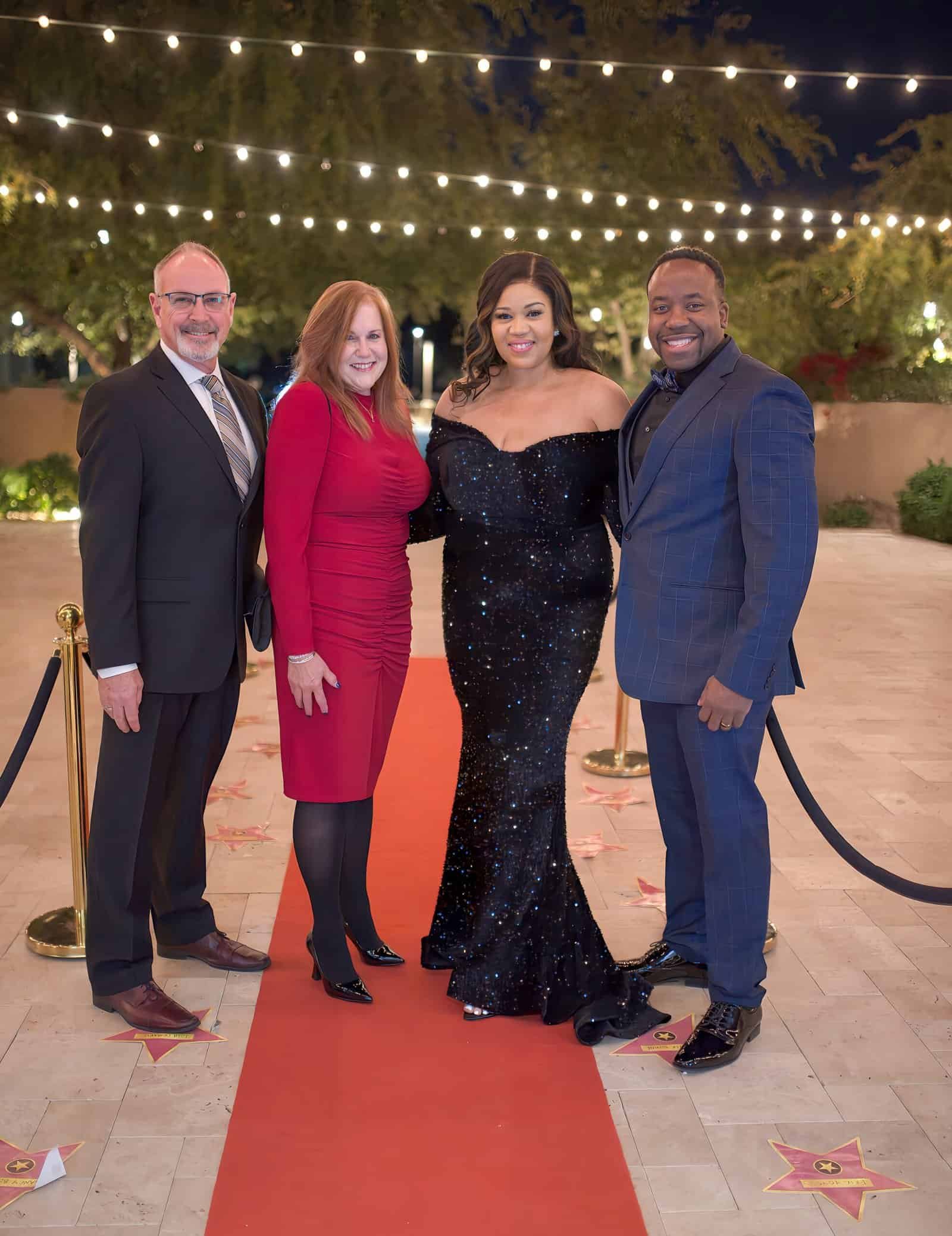 A stylish couple dressed in formal wear standing on a red carpet at an outdoor event, illuminated by string lights, capturing a special moment.