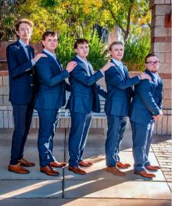 Five boys dressed in matching blue suits pose outdoors during a special occasion.