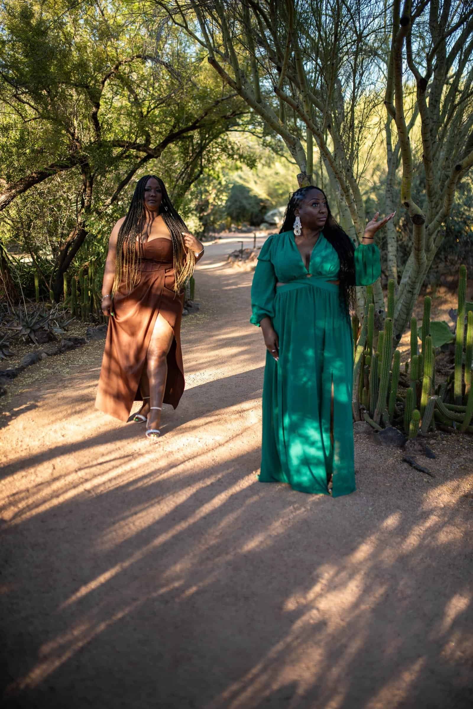 Two women dressed in stylish outfits strolling through a beautiful garden with trees and natural light.