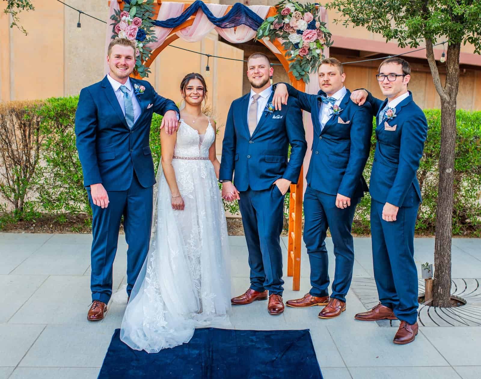 Joyful wedding moment with bride, groom, and groomsmen outdoors under floral arch.