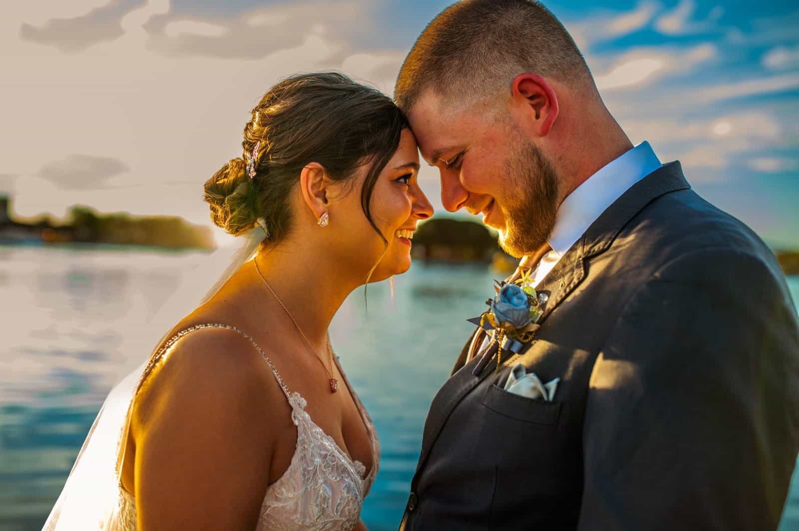 Beautiful wedding photo of a bride and groom smiling and touching foreheads during sunset by the water.