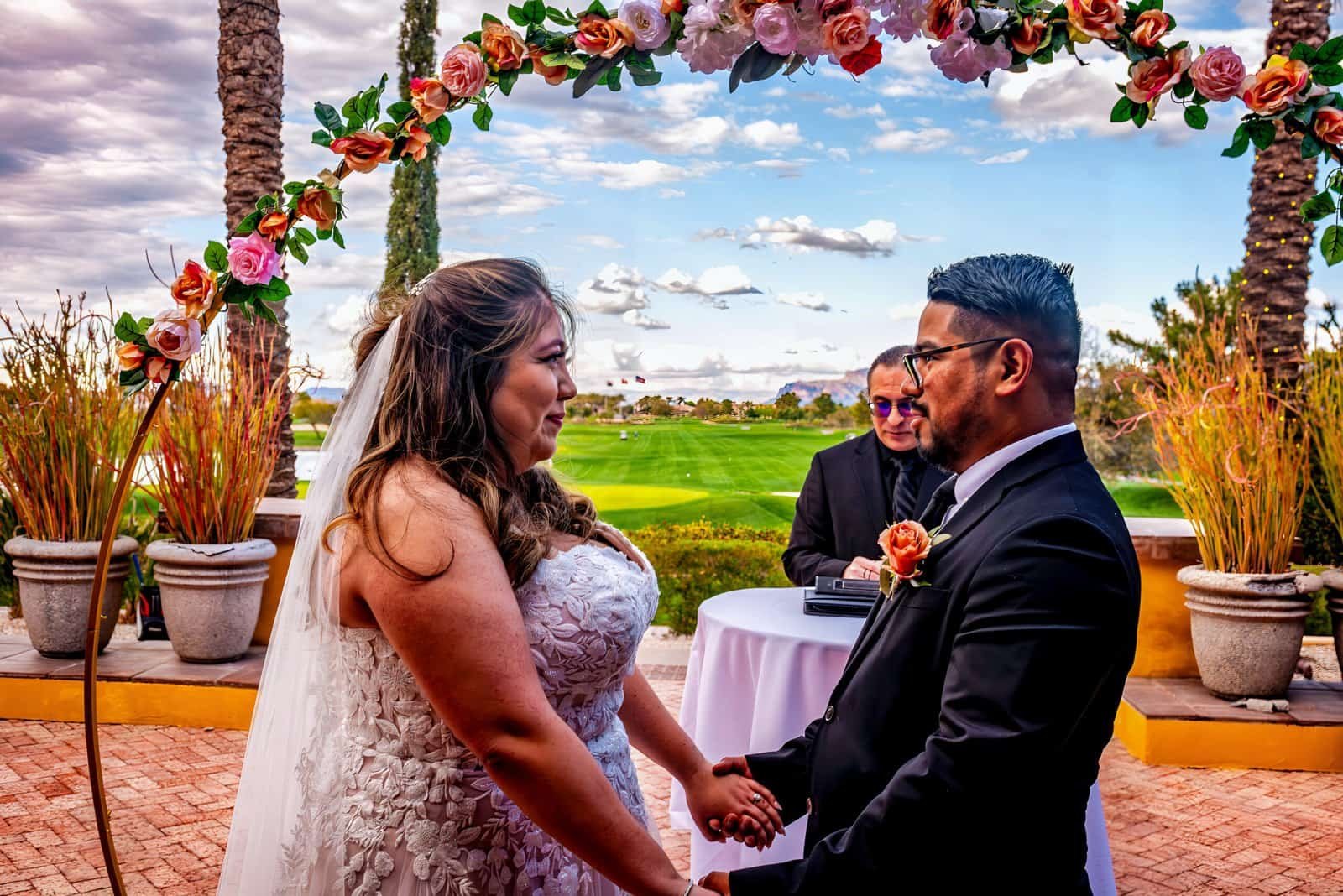 A joyful couple getting married outdoors under a decorated floral arch, capturing a special moment during their wedding celebration.