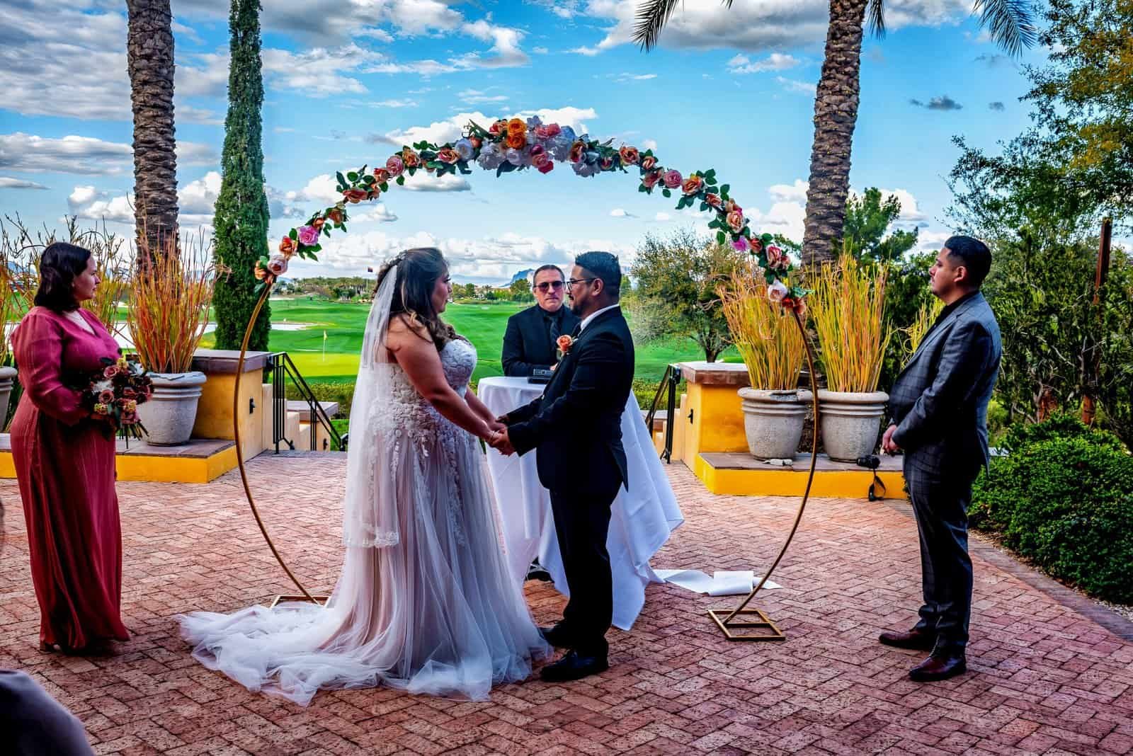 Romantic outdoor wedding ceremony with couple holding hands under a floral arch, surrounded by lush greenery and palm trees.