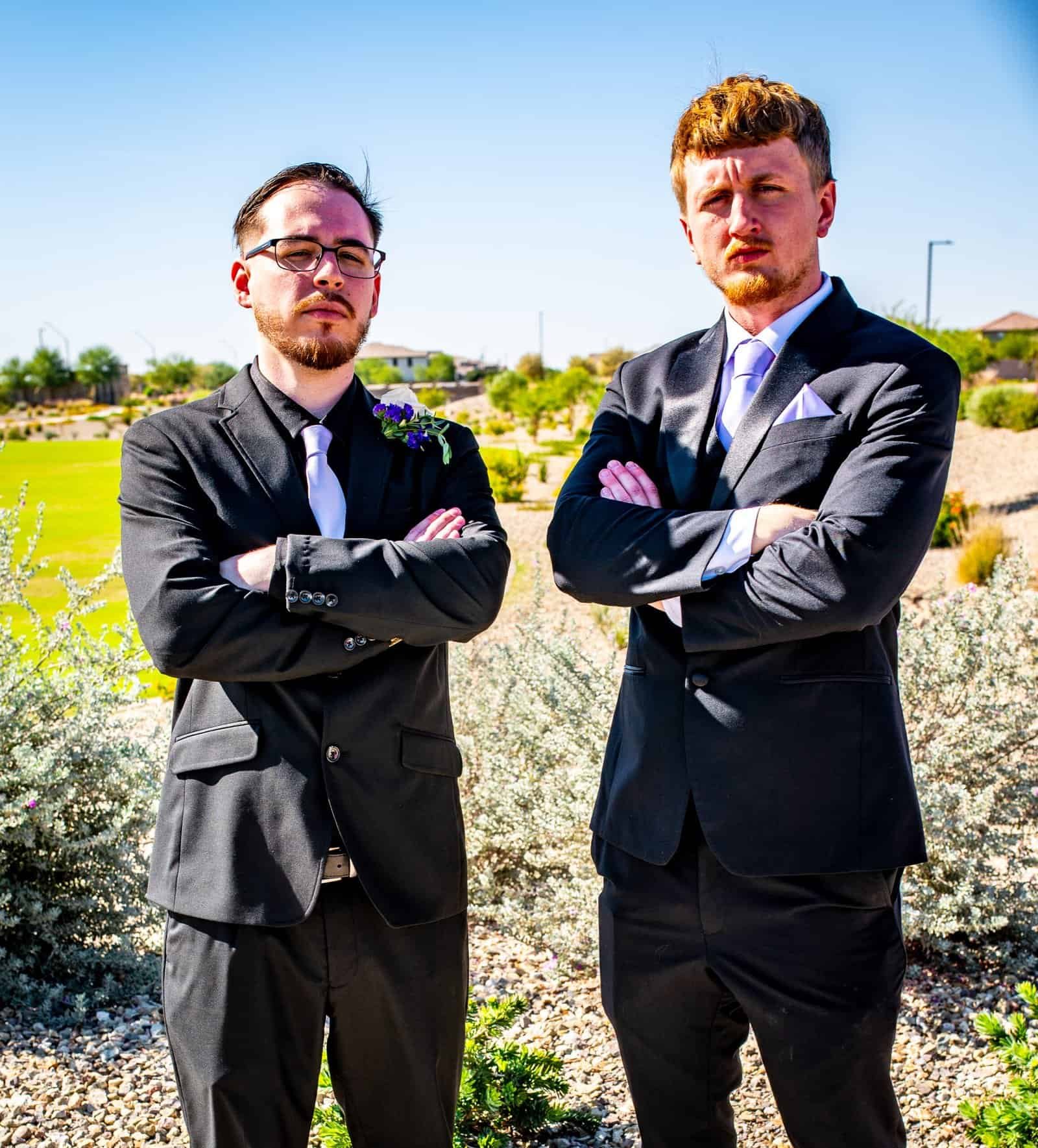 Two groomsmen in formal suits standing outdoors with arms crossed, celebrating a wedding.