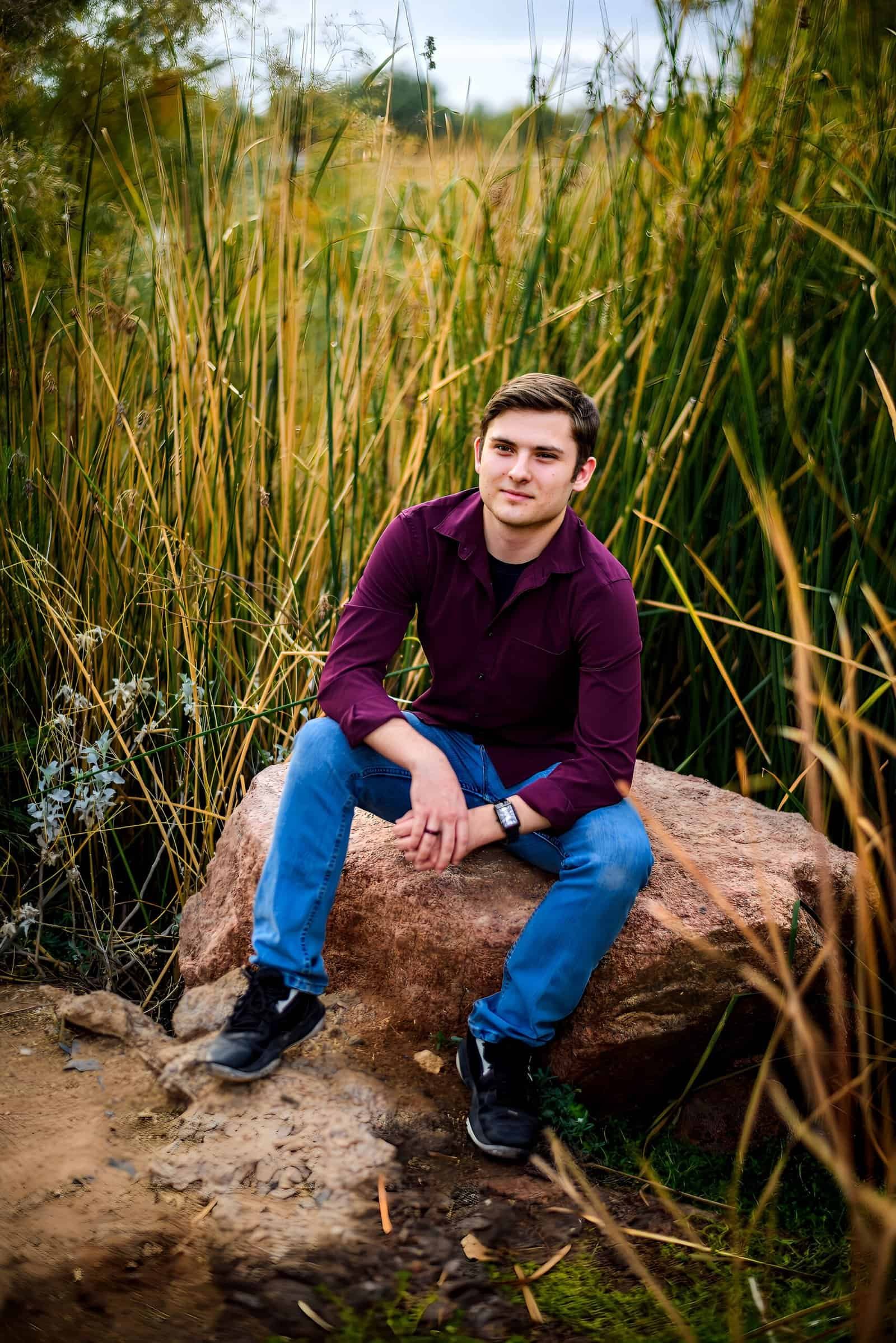 A young man in casual attire sitting on a large rock surrounded by lush greenery and tall grass, enjoying a peaceful moment outdoors.