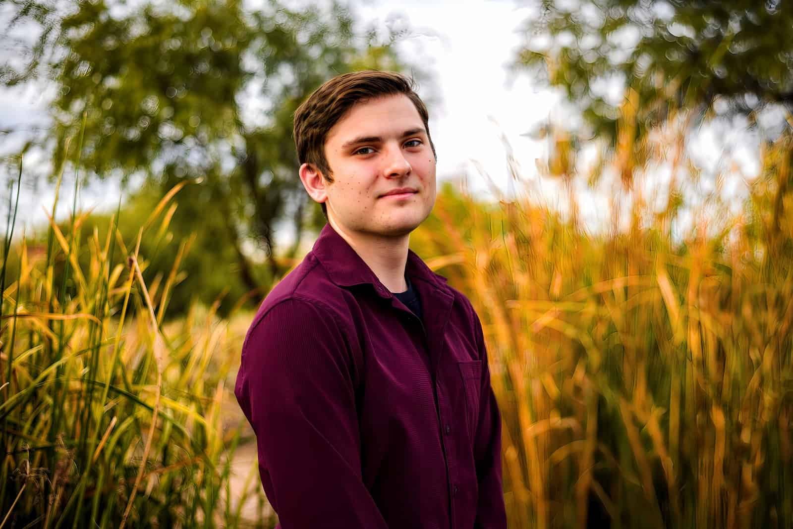 A young man in a maroon shirt posing in a scenic outdoor environment with lush greenery.