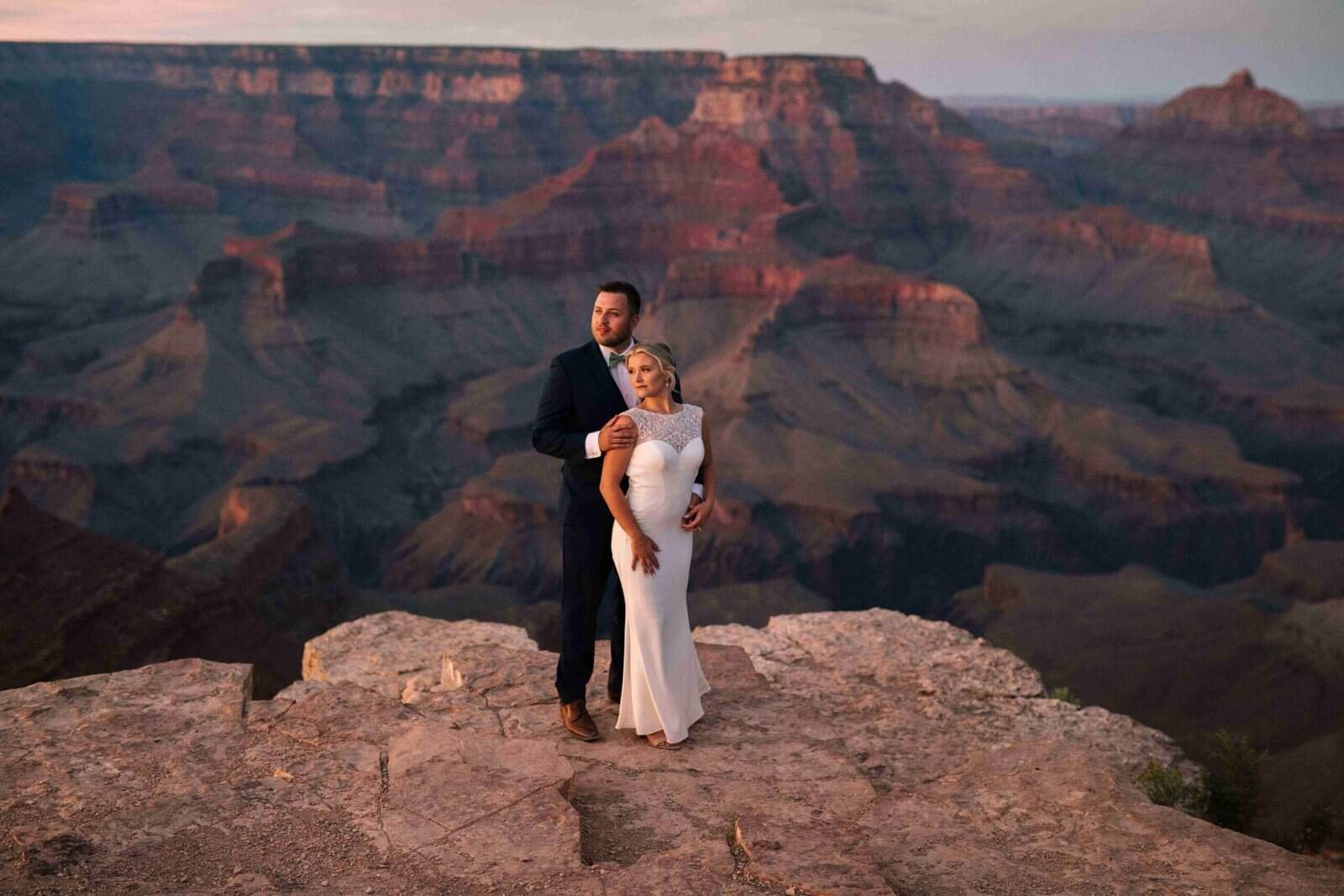 A couple in wedding attire standing on a rocky ledge overlooking the Grand Canyon during sunset, capturing a romantic moment.