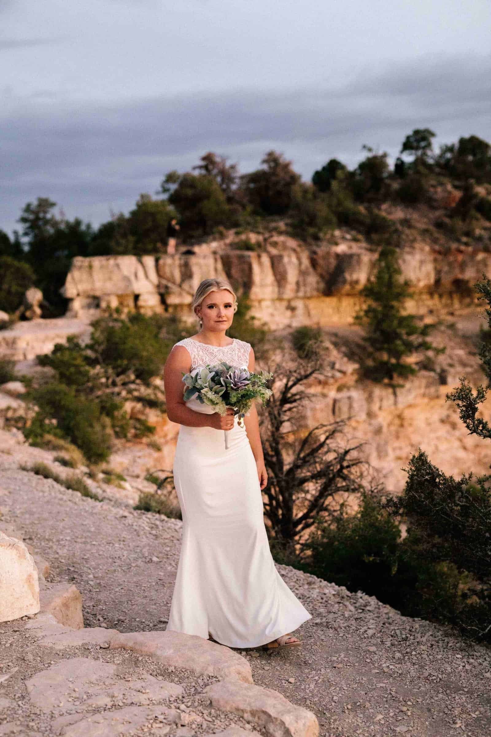 A beautiful bride in a white wedding gown standing outdoors in a rugged canyon with trees and rocky formations.