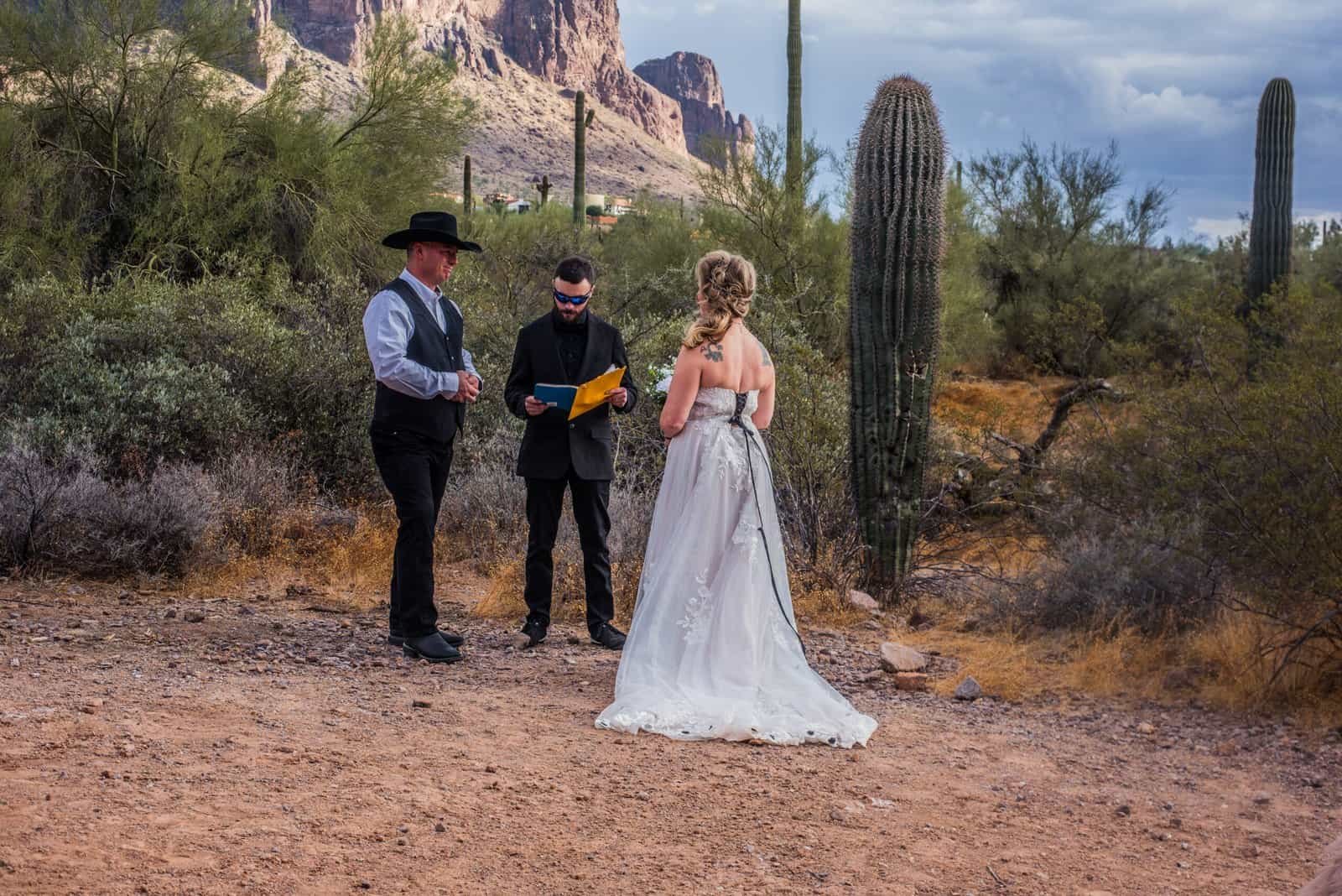 A couple gets married outdoors in a desert setting with a officiant, surrounded by cacti and scenic mountain views.