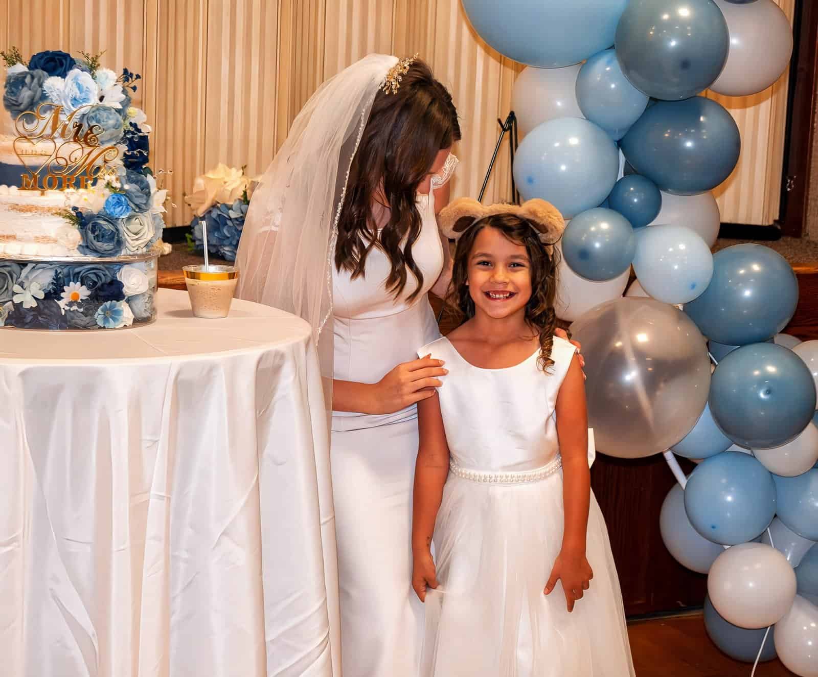 A bride and a young girl smiling together during a wedding event, surrounded by balloons and floral decor.