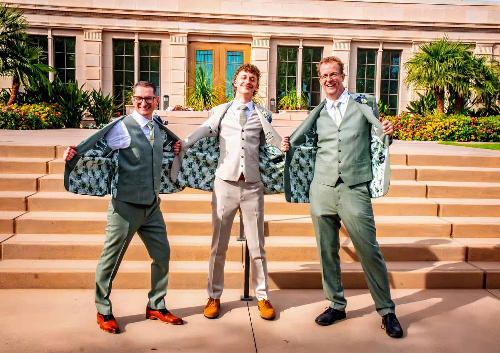 Three groomsmen in suits celebrating a wedding outdoors, holding jackets open.