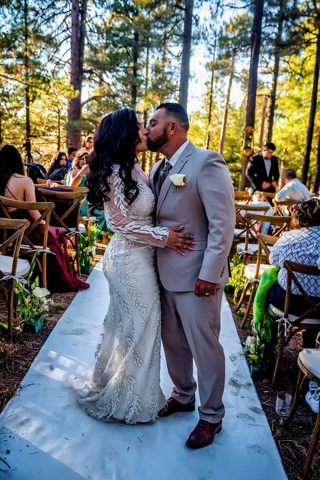 Beautiful outdoor wedding with couple kissing surrounded by guests in a forest setting.